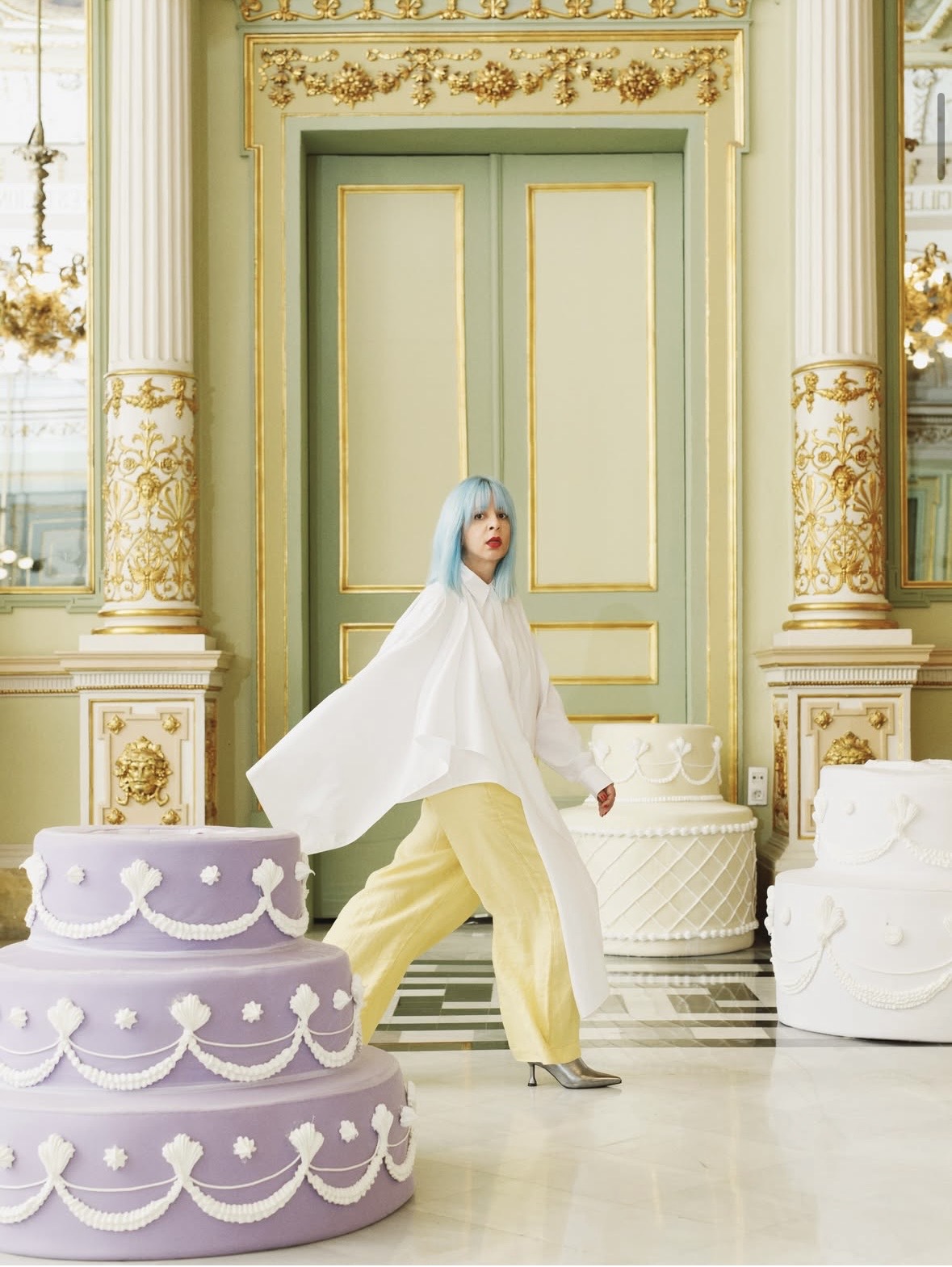 Woman with blue hair walking past giant cakes in a gilded hall